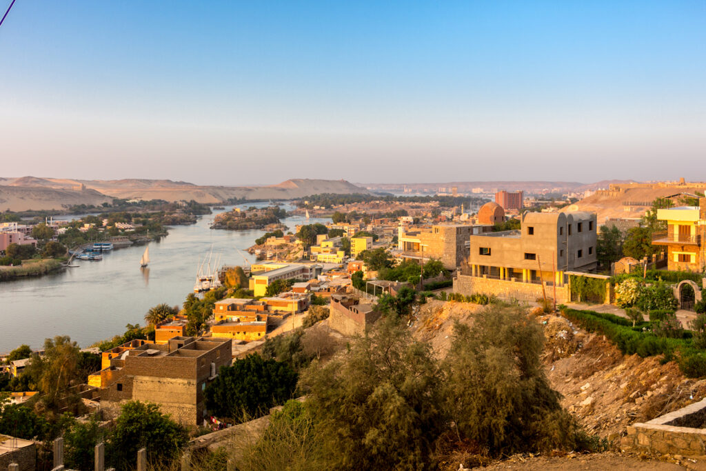 A scenic view of a riverside town with the Nile River winding through the landscape, bordered by buildings, trees, and barren hills. Several sailboats are visible on the water, and the sky is clear and blue, suggesting a calm, serene environment perfect for river cruises.