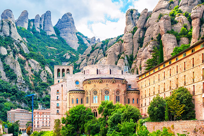 A monastery with intricate architecture nestled among towering, jagged mountains under a partially cloudy sky. The buildings are surrounded by lush greenery, and the rocky peaks form a dramatic backdrop.