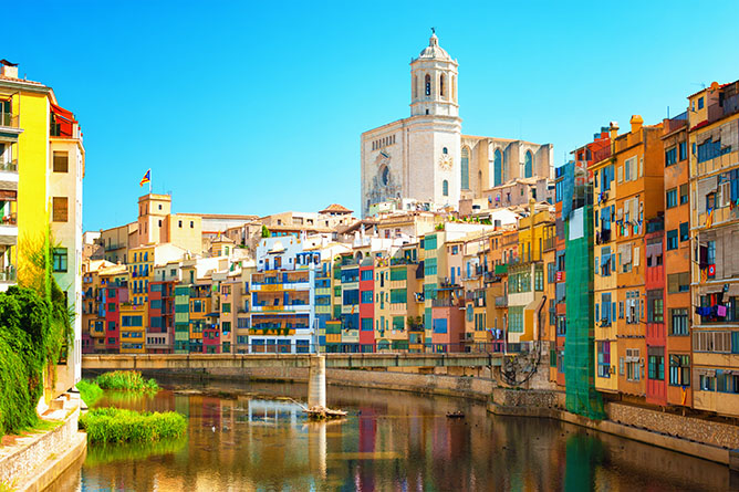 A picturesque view of colorful buildings lining the Onyar River in Girona, Spain, with the iconic Girona Cathedral towering in the background under a clear blue sky. The riverbanks are lush with greenery, adding to the vibrant scenery.