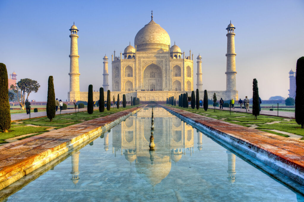 The image shows the Taj Mahal, an ivory-white marble mausoleum in Agra, India. Often referred to as the Heart of India, it features a large central dome flanked by four minarets, with a reflecting pool and symmetrical gardens in the foreground. The sky is clear, giving a serene ambiance.