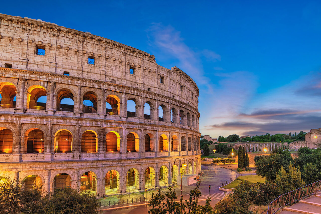 The Colosseum in Rome, Italy, illuminated at dusk. Unwrap Rome as the ancient amphitheater's arches glow with warm light against a deepening blue sky. Surrounding greenery and distant ruins are visible, highlighting the historical site's grandeur and serene ambiance.