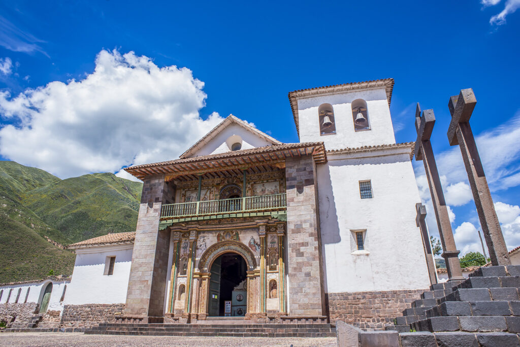 A white colonial-era church with a wooden balcony and decorative entrance stands against a bright blue sky with fluffy white clouds. To the right, three large stone crosses are positioned, and green hills serve as a backdrop.