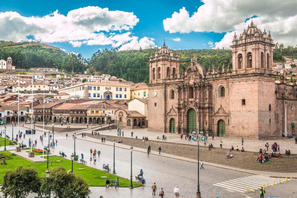 A vibrant cityscape of Cusco, Peru, showcasing the historic Plaza de Armas. The Cathedral of Santo Domingo stands prominently with its ornate facade. The square is lively with pedestrians, greenery, and surrounding colonial buildings under a partly cloudy sky—an ideal spot for Peru vacation packages with airfare.