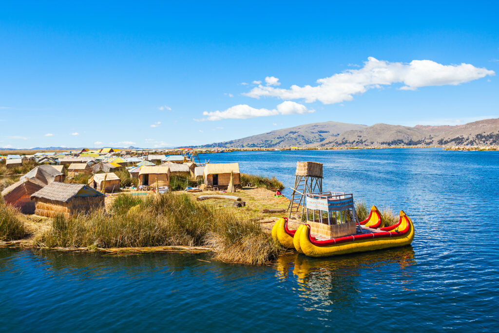 A scenic view of a floating island on Lake Titicaca, with traditional thatched huts and reed boats under a bright blue sky. The surrounding landscape includes mountain ridges and vast waters, with clouds scattered across.