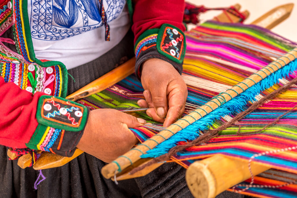 Close-up of a person weaving colorful fabric on a traditional loom. The weaver is wearing a red garment adorned with intricate embroidery. Brightly colored threads are being woven into a pattern, showcasing vivid blues, greens, pinks, and yellows.