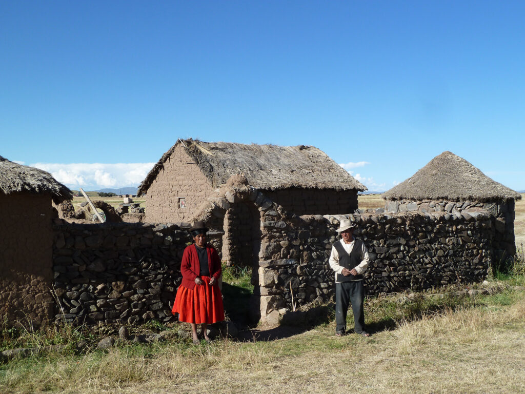 Two people stand outside traditional stone and thatch buildings in a rural area. One person is wearing a red jacket and dress, while the other is dressed in dark pants and a vest with a white shirt and hat. The sky above is clear and blue.