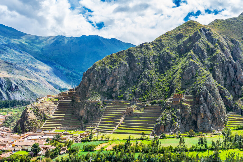 A scenic view of an ancient site in Ollantaytambo, Peru, featuring terraced stone structures built into the hillside. The green terraces contrast with the rocky mountain and the lush valley below, all set against a backdrop of distant mountains under a partly cloudy sky.