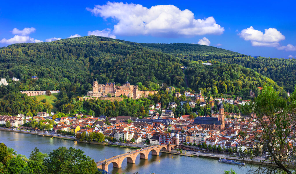 Scenic view of Heidelberg, Germany, featuring the Old Bridge (Karl Theodor Bridge) crossing the Neckar River, the historical Heidelberg Castle perched on the hillside, and a backdrop of lush green hills under a vibrant blue sky with scattered clouds—a perfect destination for those exploring Rhine river cruises from Amsterdam.