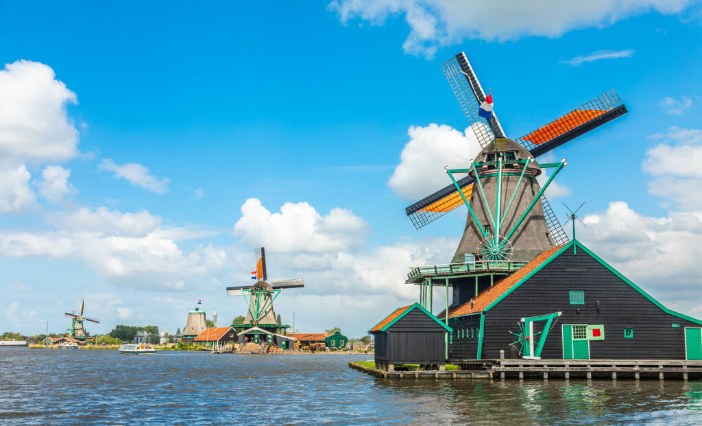 Experience the scenic view of traditional Dutch windmills along a calm river under a blue sky with scattered clouds. The windmills, with their large wooden blades, stand among a green, picturesque landscape, showcasing historic architecture with vibrant colors—a perfect sight on Rhine River cruises from Amsterdam.