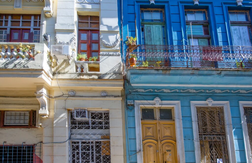 Colorful facade of two adjacent buildings, one white and one blue, in captivating Cuba. Both buildings feature ornate architectural details with balconies adorned with potted plants and large wooden doors. Sunlight casts shadows, highlighting the intricate designs and vibrant colors.