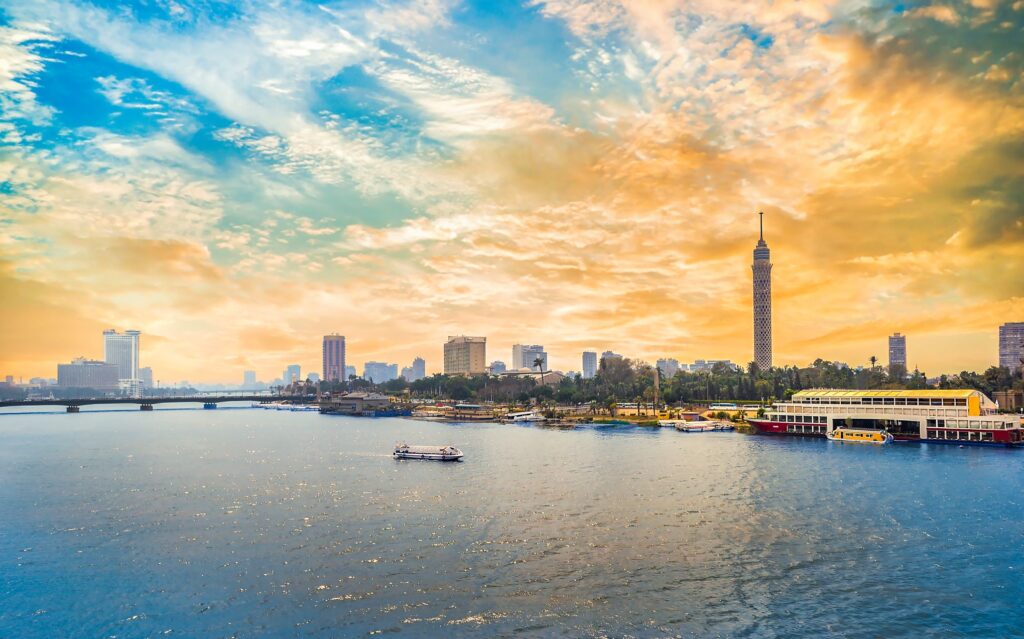 A riverside cityscape at sunset, featuring the Cairo Tower on the right, high-rise buildings in the background, a bridge on the left, and boats on the Nile River in the foreground. The sky is painted in warm hues of orange, pink, and blue with scattered clouds—a perfect highlight for Egypt tour packages.