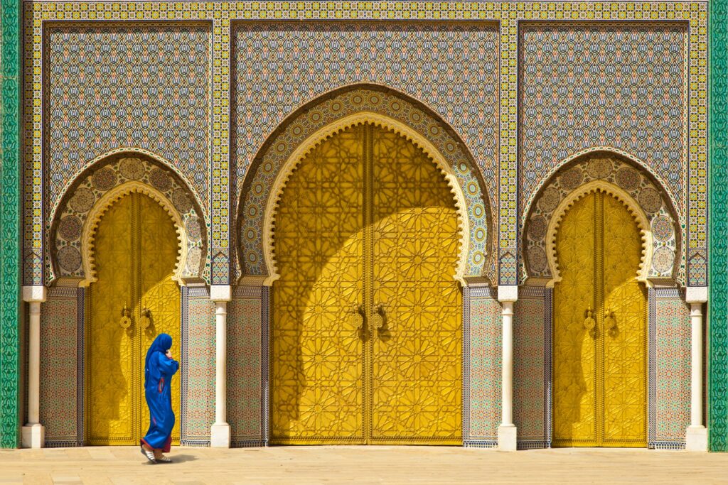 A person wearing a blue traditional garment walks in front of large, ornate golden doors with intricate patterns and arched frames. The doors, often admired on Morocco escorted tours, are set in a decorative, tiled wall with a mosaic design.