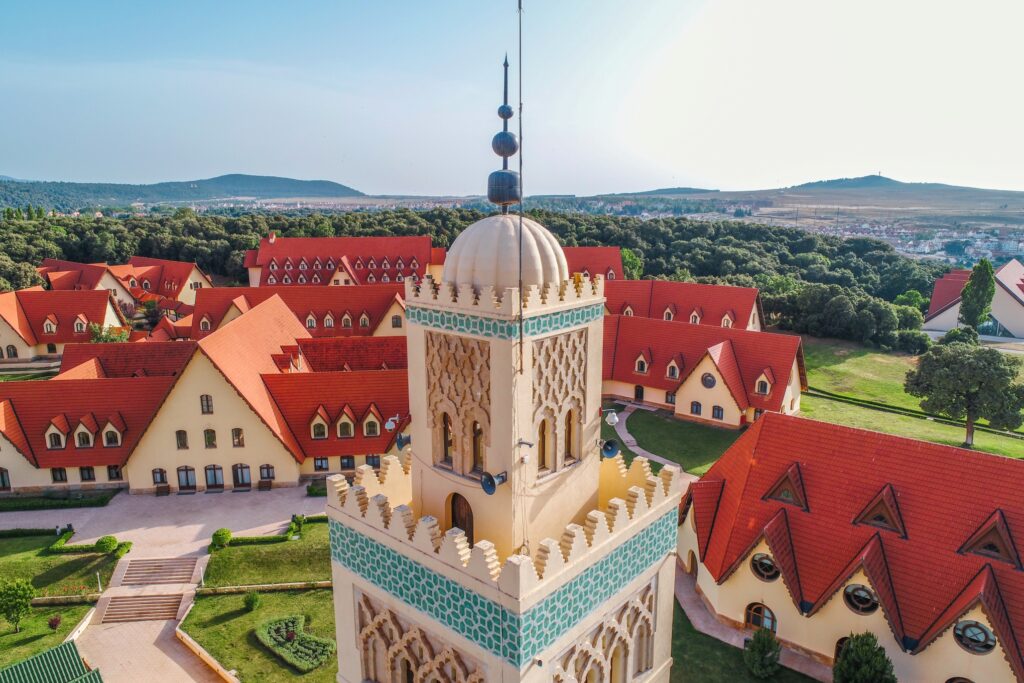 An aerial view of a picturesque village with numerous buildings featuring red-tiled roofs. In the foreground is a prominent tower with detailed carvings and green accents. Rolling hills and greenery surround the village under a clear blue sky, reminiscent of scenes from Morocco Escorted Tours.