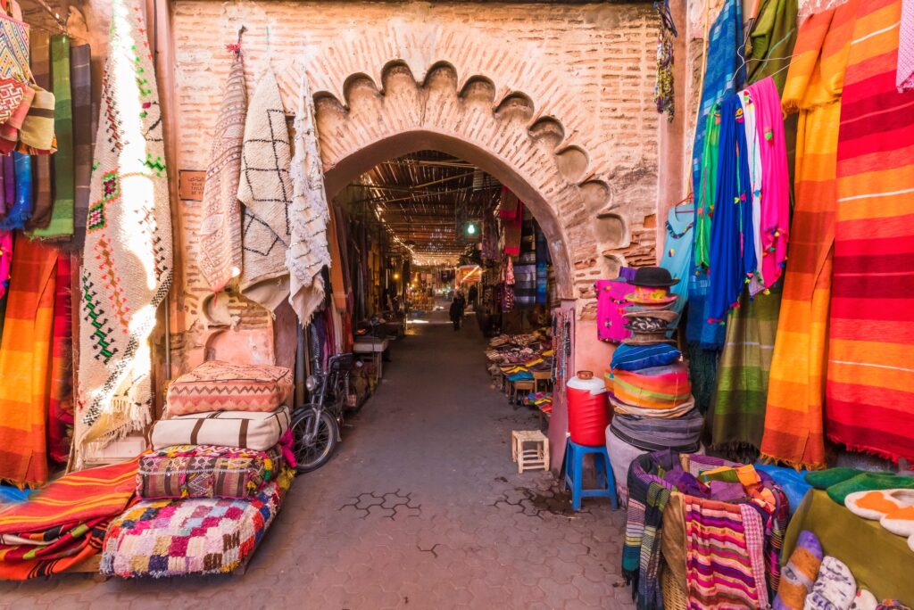 A vibrant market with colorful textiles, blankets, and rugs displayed on either side. The street is narrow, leading through an arched brick gateway. Various items are stacked neatly, creating a rich tapestry of colors and textures. On one of the Morocco Escorted Tours, you'll find a bicycle leaning against the left side.