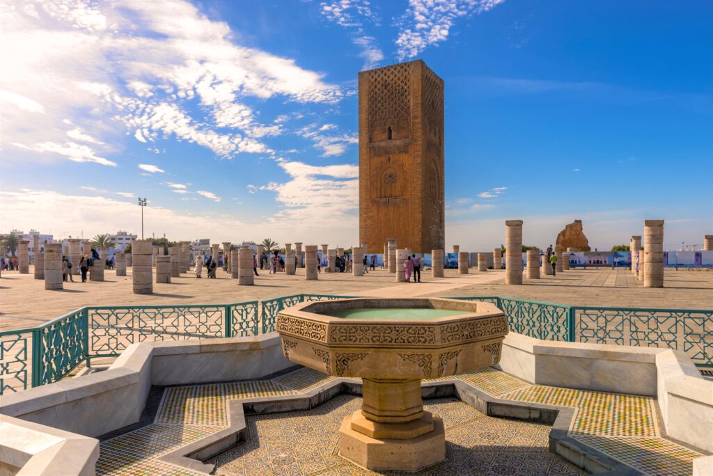 A historic site with a large, intricate stone tower under a clear blue sky, surrounded by numerous pillars and visitors. In the foreground, there is an ornate fountain with colorful mosaic tiles and a decorative green railing—ideal for exploration on Morocco Escorted Tours.