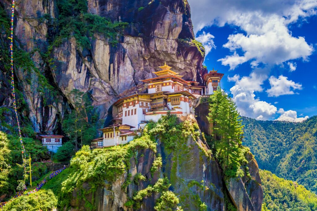 A beautiful monastery, Paro Taktsang or Tiger's Nest, clings to a cliffside amid lush greenery in the Himalayan region of Bhutan. As one of the wonders of the East, its traditional design contrasts with the rugged, forested mountain backdrop under a bright blue sky dotted with white clouds.