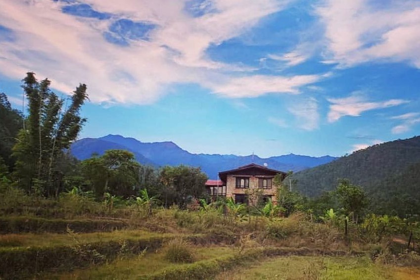 A wooden house is nestled among lush greenery with Himalayan mountains in the background. The sky is scattered with wispy clouds. Bamboolike trees and various plants surround the area, creating a serene and picturesque landscape full of natural wonders.