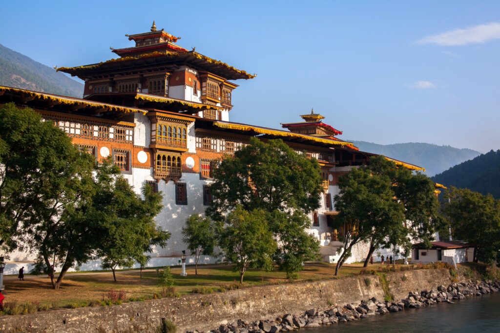 A grand traditional Bhutanese fortress sits majestically by the riverside, with its white walls and ornate wooden windows. Surrounded by lush trees and set against a backdrop of rolling Himalayan hills under a clear blue sky, this wonder exudes historical and cultural significance.