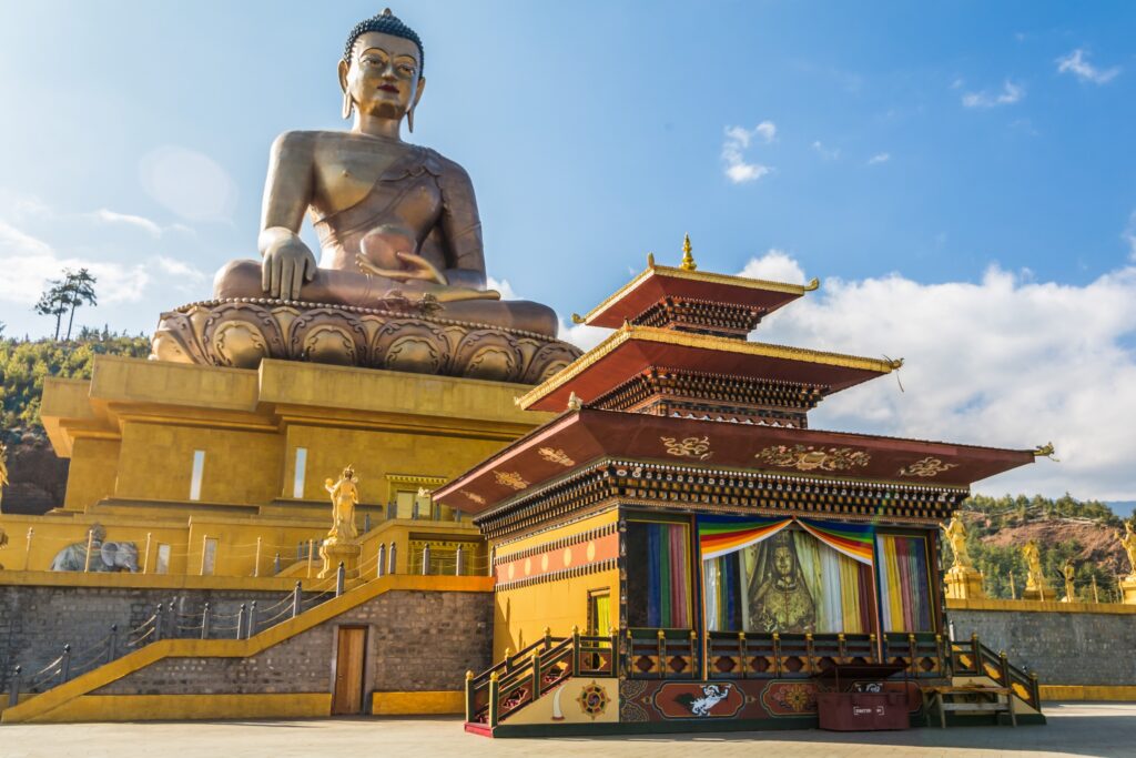 A large golden Buddha statue sits atop a high pedestal under a blue sky with scattered clouds. In the foreground, a smaller, ornately decorated structure with colorful prayer flags is visible, embodying the Himalayan wonders. The setting appears serene with nature in the background.