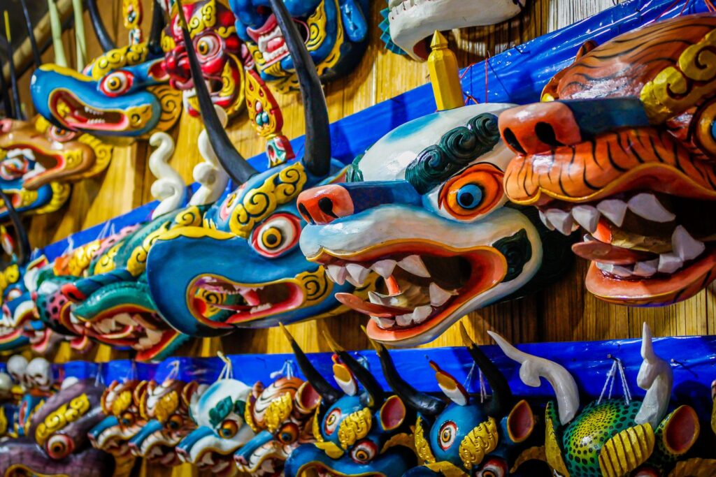 A vivid display of intricately painted traditional masks, featuring dragon, tiger, and demon designs with vibrant colors and exaggerated expressions. The masks are arranged closely together on a wooden wall, highlighting detailed craftsmanship and the cultural artistry of Himalayan wonders.