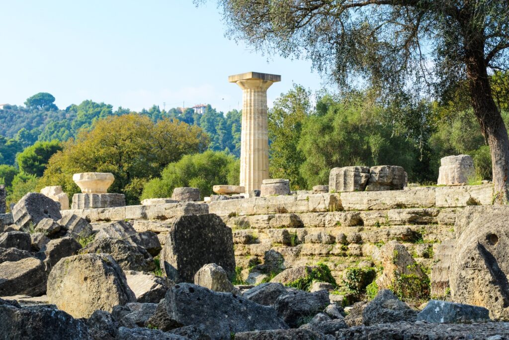 Ruin site featuring a solitary ancient column standing amidst scattered stone blocks and fragments. The scene, reminiscent of spectacular Greece, is surrounded by greenery, including trees and distant hills under a clear blue sky.