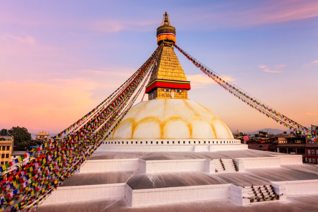 A majestic white stupa with a golden spire and colorful prayer flags stands tall against a vibrant, pastel-colored sunset sky. Its watchful eyes are reminiscent of the awe-inspiring Wonders of the Himalayan region. Surrounding buildings accentuate its tranquil beauty.