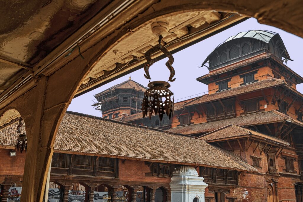 The image showcases an architectural view of traditional Nepali buildings made of red bricks and wooden elements. The structures, adorned with intricate wooden carvings, stand as Himalayan wonders seen through an arched, decorated doorway. A lantern hanging from the archway is in the foreground.