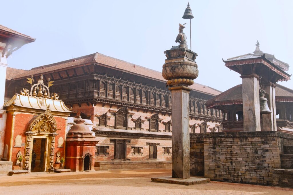 A photo of a historic square featuring traditional South Asian architecture, including a large ornately decorated gate on the left, a tall column with a statue on top, and a pagoda-style pavilion to the right. The backdrop includes an elaborate multi-storied building evoking Himalayan wonders.