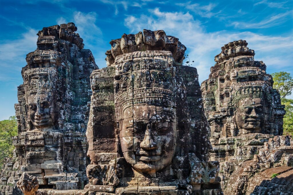 Stone faces of Bayon Temple in Angkor Thom, Cambodia. The ancient, intricately carved faces are part of a massive sandstone structure surrounded by lush greenery under a bright blue sky with scattered clouds. The central face is prominently featured with serene expression, perfect for Thailand and Angkor Wat tours.