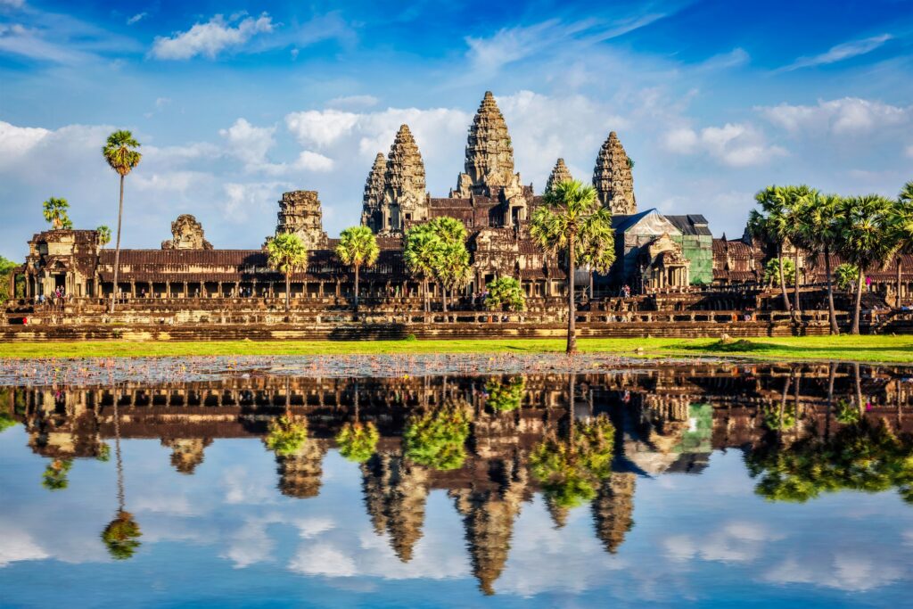 A panoramic view of Angkor Wat in Cambodia with its distinctive towers and intricate stone architecture. The temple complex, often featured in Thailand and Angkor Wat tours, is reflected in a calm body of water in the foreground, surrounded by lush greenery under a vibrant blue sky with scattered clouds.