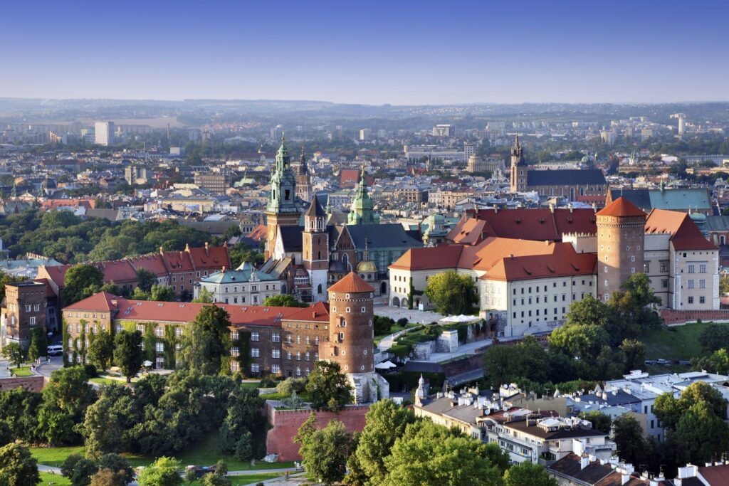 Aerial view of Wawel Castle in Kraków, Poland, featuring red-roofed buildings surrounded by lush greenery. The cityscape stretches into the distance under a clear blue sky, showcasing both historical architecture and modern structures—perfect for adding to a Prague or Vienna tour itinerary.