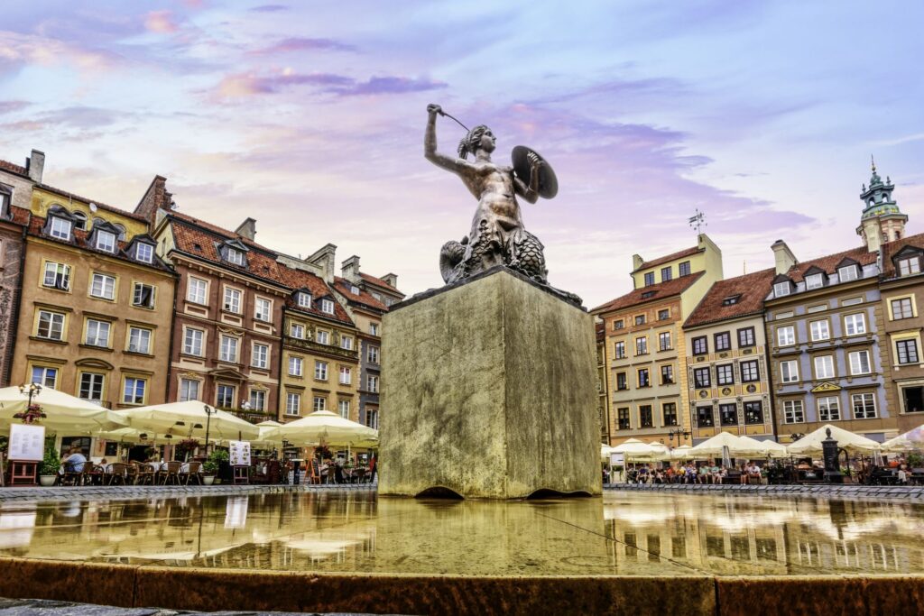 A statue of a mermaid holding a sword and shield stands atop a pedestal, surrounded by water in a fountain. The background features historic buildings with red-tiled roofs and numerous outdoor seating areas under umbrellas, reminiscent of strolls through Prague or Vienna. The sky is colored with hues of purple and orange.