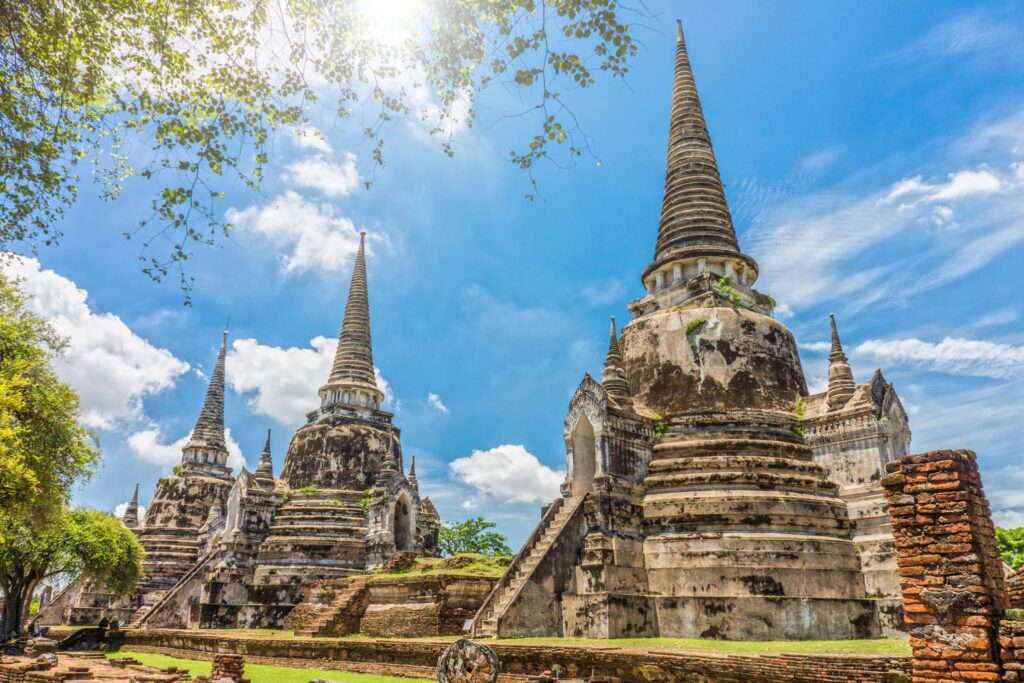 Ancient stone temples with tall, pointed spires at Wat Phra Si Sanphet in Ayutthaya, Thailand, under a bright blue sky with scattered clouds. The overgrown trees and sunlit surroundings embody the spirit of Thailand, creating a serene atmosphere.