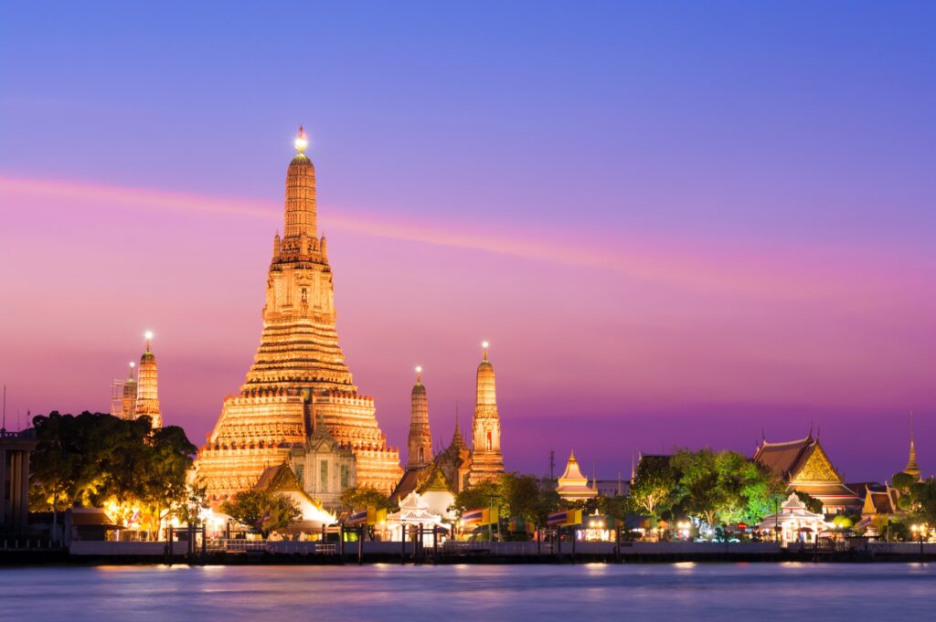 A stunning view of Wat Arun (Temple of Dawn) in Bangkok, Thailand, illuminated at twilight. The temple's spires are glowing under a vibrant pink and purple sky, with reflections visible in the calm water of the Chao Phraya River in the foreground—a true travel spirit encapsulated.