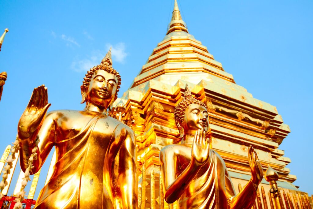 Two golden Buddha statues stand in front of an ornate, gold-colored pagoda against a clear blue sky. The statues are intricately detailed, with one statue showing a raised right hand in a blessing gesture. The scene conveys the spirit of Thailand, exuding peace and reverence.