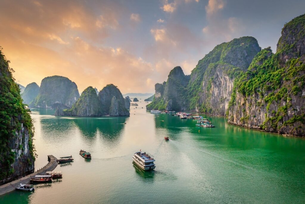 A serene view of Halong Bay during sunset, featuring numerous limestone karsts and islets. Several boats float on the calm emerald green water, and lush vegetation covers the rocky cliffs. Luxurious cruise ships and smaller boats dock near the shore, showcasing top Vietnam attractions.