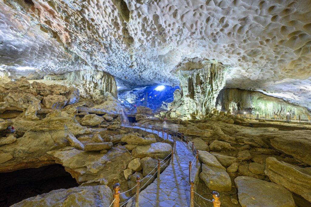 A vast cave interior with a rocky pathway flanked by ropes leads deeper into the cavern. The cave walls and ceiling are textured with calcite formations, illuminated by artificial lighting that creates a mix of white and turquoise hues—a true gem among Vietnam attractions.