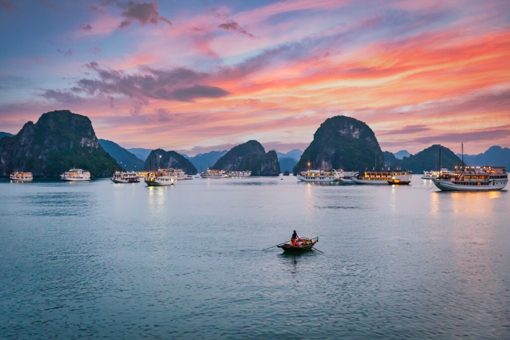 A tranquil scene at sunset features a single person rowing a boat on calm waters surrounded by larger boats and limestone karsts under a colorful sky with hues of pink, purple, and orange—truly one of the Vietnam highlights that makes Vietnam tourism so enchanting.