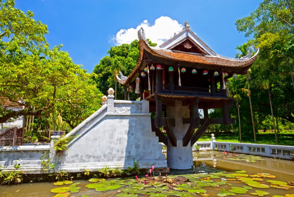 Image of the One Pillar Pagoda in Hanoi, a historic Buddhist temple and one of the Vietnam attractions. The pagoda stands on a single stone pillar amidst a pond filled with lily pads and flowers. Surrounding the structure are lush green trees under a bright blue sky with a few white clouds.