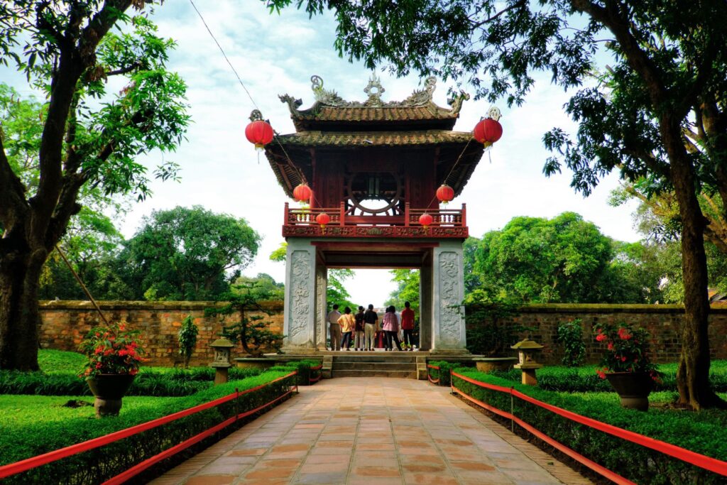 A serene pathway leads to the Temple of Literature, a historic highlight with traditional architecture in Hanoi, Vietnam. The temple features a pagoda-style roof adorned with red lanterns. Lush greenery surrounds the pathway, and a group of people stands at the entrance.