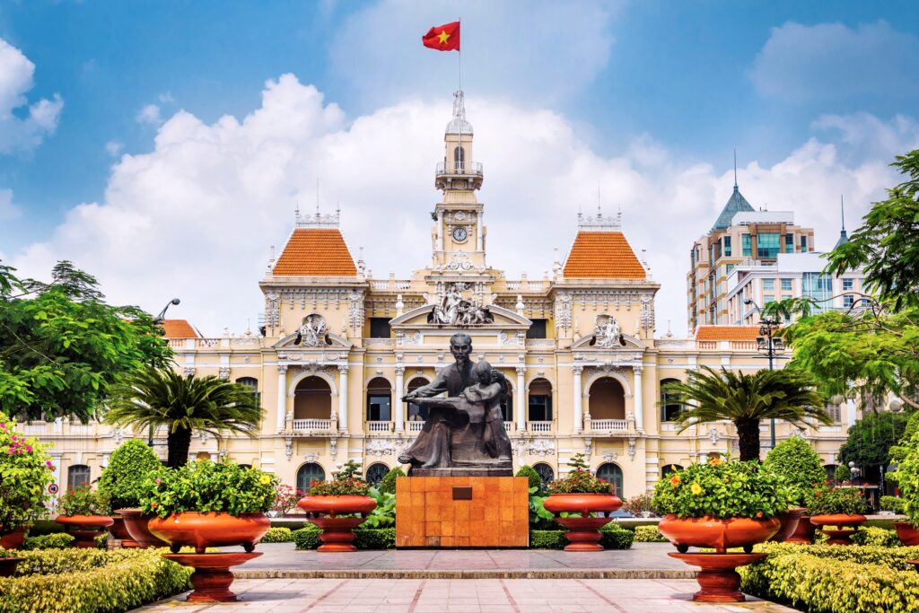 A picturesque view of Ho Chi Minh City Hall, a highlight of Vietnam tourism. The colonial-style building features a large central clock tower and is topped with the Vietnamese flag. The foreground includes a statue surrounded by neatly arranged flower pots and lush greenery, making it one of the top Vietnam attractions.