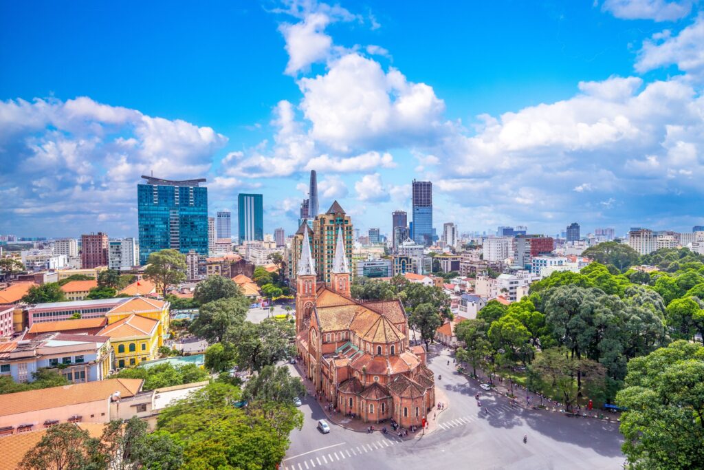 Aerial view of a cityscape featuring a historic red-brick cathedral with twin bell towers in the foreground surrounded by trees. Modern skyscrapers and various buildings are visible in the background under a bright blue sky with scattered clouds, showcasing Vietnam highlights.