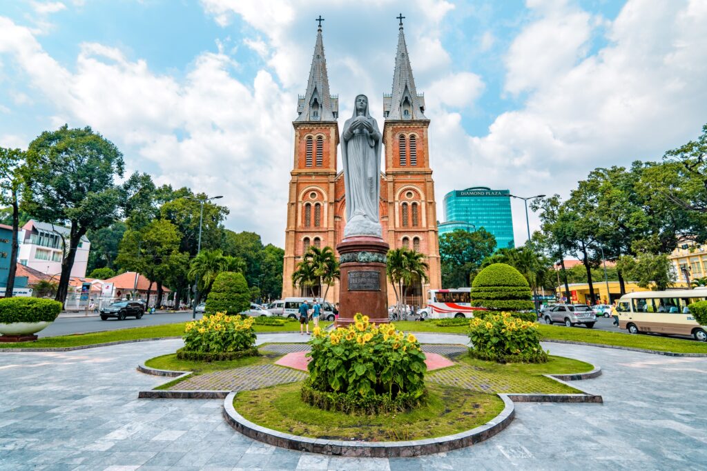 A statue of the Virgin Mary stands in front of the Notre-Dame Cathedral Basilica of Saigon, one of the top Vietnam attractions in Ho Chi Minh City. The cathedral, a highlight of Vietnamese culture, features two towering spires and a brick facade, set against a well-maintained garden and blue sky with clouds.