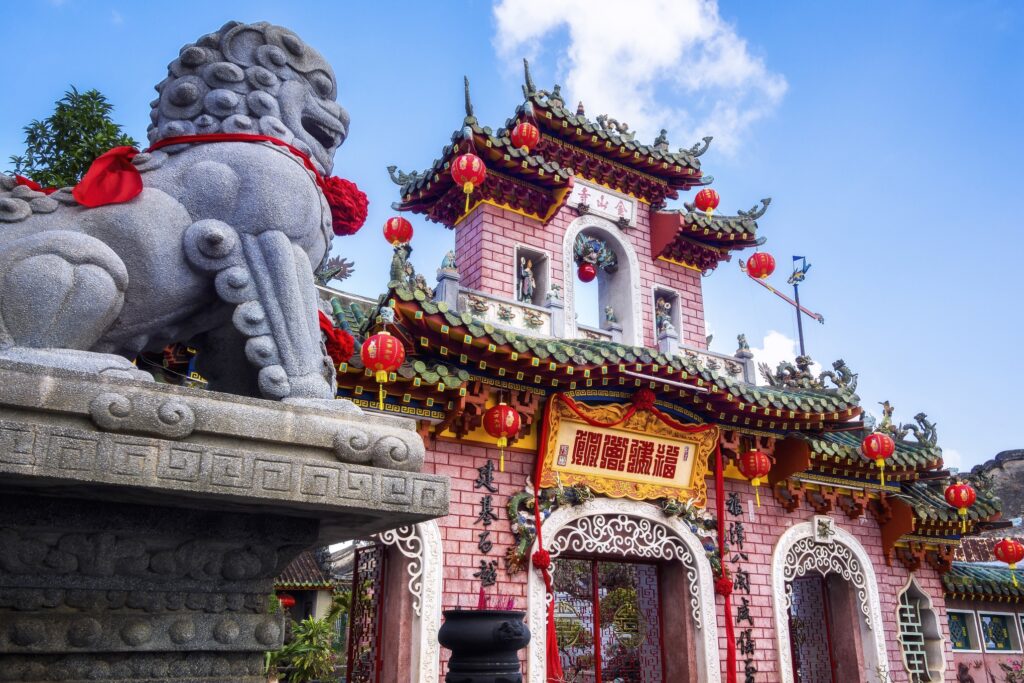 A vibrant pink Chinese temple with ornate architecture and red lanterns hanging around. In the foreground, a stone lion statue adorned with a red ribbon stands proudly. The temple's entrance features intricate designs and Chinese characters on banners. The bright blue sky completes this Vietnam tourism gem.