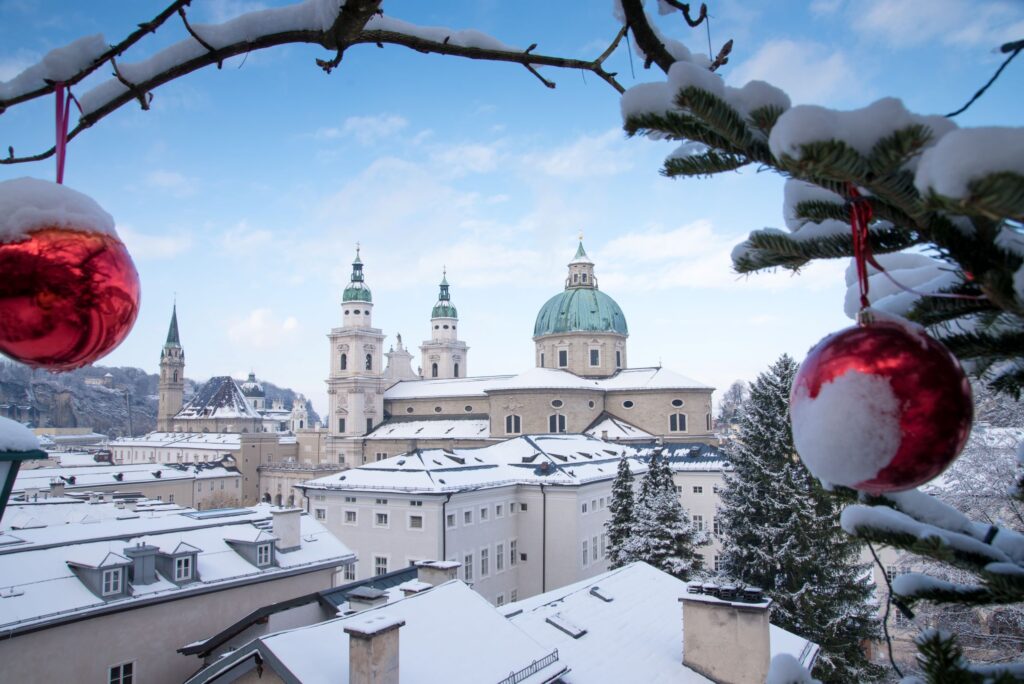 A scenic winter view of Salzburg, Austria, featuring the Salzburg Cathedral surrounded by snow-covered buildings. The foreground includes snow-covered tree branches decorated with red baubles, evoking a charming Christmas atmosphere. This picturesque cityscape unfolds under a clear blue sky.