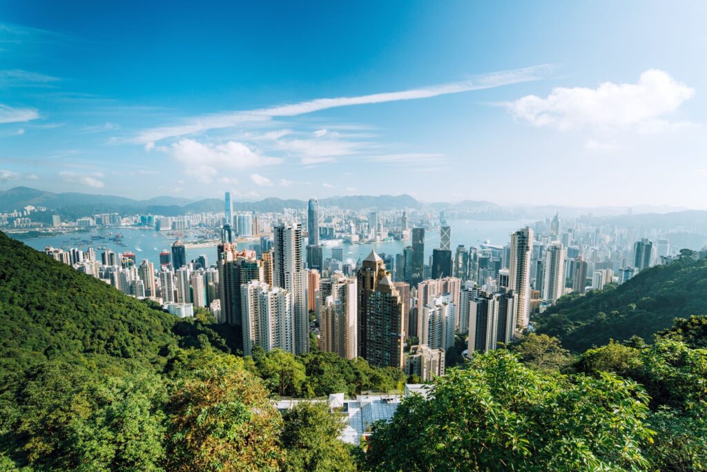 View of a densely built cityscape featuring tall skyscrapers against a backdrop of a blue sky and distant mountains. The foreground includes lush green trees, while a harbor is visible in the middle distance. The scene brings to mind the urban landscape of Hong Kong surrounded by natural beauty.