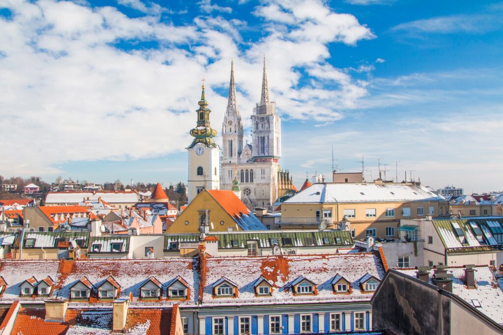 Rooftop view of Zagreb with light snow covering the buildings. In the center, Zagreb Cathedral with its twin spires rises prominently against a backdrop of a partly cloudy blue sky. Surrounding buildings display colorful facades and classic Old World architecture, evoking a festive Christmas charm.