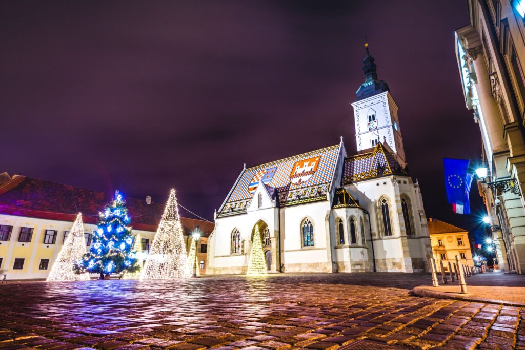 A nighttime view of a historical church with a colorful tiled roof, illuminated by festive lights. A decorated Christmas tree and lit cone-shaped decorations stand nearby, evoking an Old World charm. The surrounding buildings and cobblestone street add to the festive atmosphere reminiscent of traditional Christmas markets.