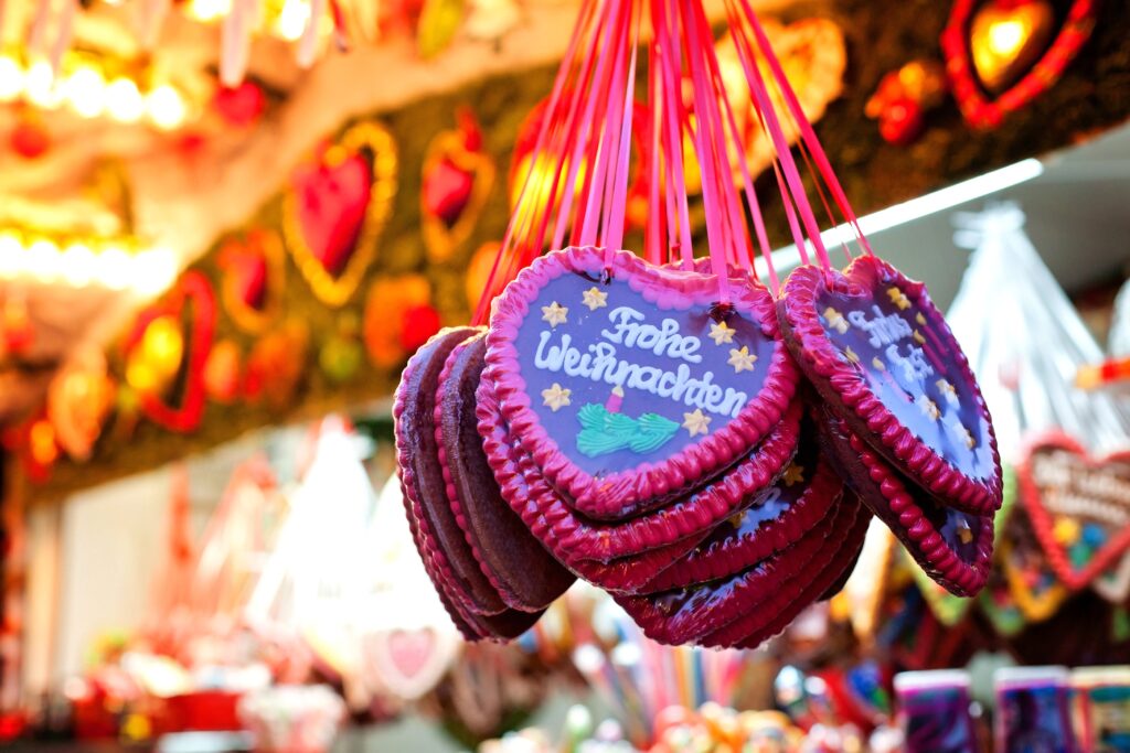 A close-up of heart-shaped gingerbread cookies hanging by red ribbons at an Old World Christmas market. The cookies are decorated with colorful icing and text that reads "Frohe Weihnachten" (Merry Christmas in German). The background features more decorated cookies and festive lights.