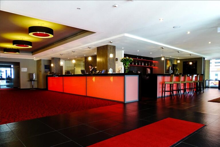 The modern hotel lobby, dressed in a festive black and red color scheme, exudes a subtle Christmas charm. Glowing red panels illuminate the reception desk, while high-backed chairs at the bar counter invite guests to unwind. A vibrant red carpet guides visitors through this contemporary winter wonderland.